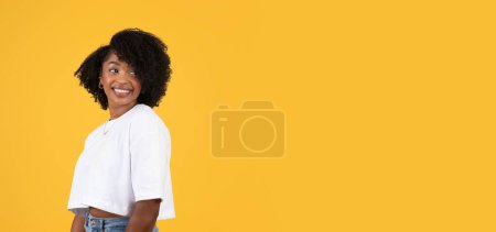Téléchargez les photos : Glad jolie jeune dame bouclée noire en t-shirt blanc regarde en arrière, bénéficie de temps libre isolé sur fond orange, studio, panorama. Vente, mode de vie, publicité et offre, émotions humaines - en image libre de droit