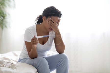 Photo for Unwanted Pregnancy Concept. Desperate young black woman in pajamas sitting alone on bed at home, african american female holding positive pregnancy test and touching her head, crying, copy space - Royalty Free Image