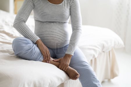 Photo for Pregnancy and health problems. Unrecognizable black pregnant woman sitting on bed at home and massaging her swollen foot, cropped shot of expectant african american mother rubbing painful ankle - Royalty Free Image
