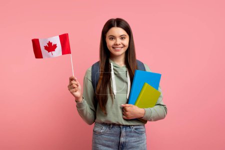Foto de Estudia en el extranjero. Hermosa chica adolescente sosteniendo la bandera de Canadá y libros de trabajo, sonriente Fejalá adolescente con mochila disfrutando de programas de intercambio de estudiantes y escuelas de idiomas, de pie sobre fondo rosa - Imagen libre de derechos