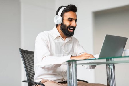 Photo for Deadline concept. Smiling indian man programmer networking and connecting to internet using laptop and wearing headphones, sitting at desk in office - Royalty Free Image