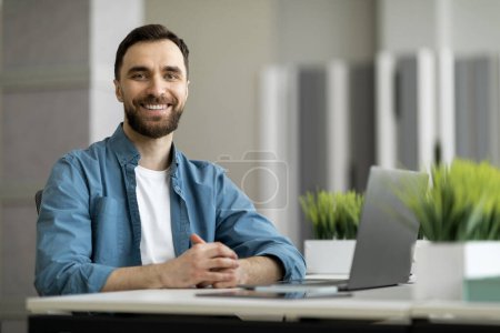 Téléchargez les photos : Portrait d'un homme indépendant assis au bureau avec ordinateur portable au bureau, homme entrepreneur heureux regardant et souriant à la caméra, appréciant le travail à distance et le travail indépendant, espace de copie - en image libre de droit