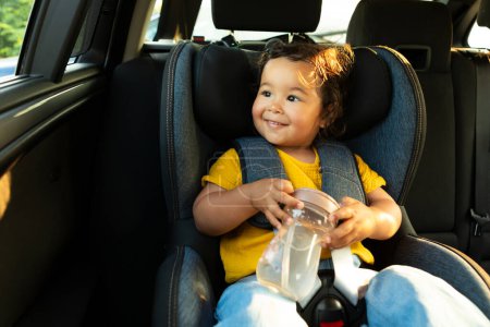 Téléchargez les photos : Voiture sûre et heureuse voyage avec bébé. Doux bébé japonais fille tenant le biberon solidement assis dans la chaise de véhicule pour tout-petit avec sangles de sécurité attachées, assis avec confort dans l'automobile moderne - en image libre de droit