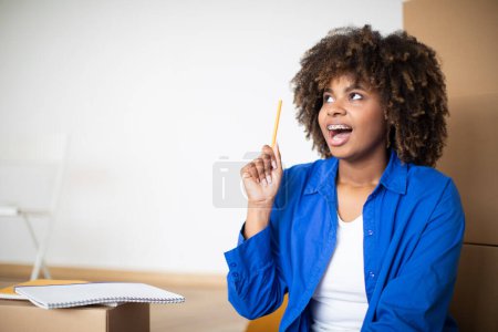 Foto de Emocionado joven mujer negra que tiene idea al hacer la lista de verificación en el día de la mudanza, alegre mujer afroamericana levantando lápiz y mirando al costado en el espacio de copia, sentado entre cajas de cartón en casa - Imagen libre de derechos