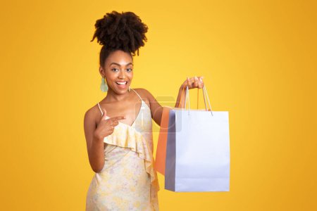 Foto de Alegre millennial dama negra adicta a las compras, disfruta de ir de compras en tiempo libre, señala con el dedo las bolsas con las compras, aislado en el fondo naranja, estudio. Anuncio y oferta, recomendación de venta - Imagen libre de derechos