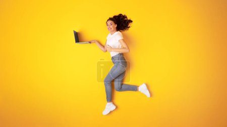 Téléchargez les photos : J'aime l'offre en ligne. Happy Young Woman tenant ordinateur portable en cours d'exécution et gestuelle pouces vers le haut, approuvant le site Web avec comme tout en naviguant sur Internet sur fond jaune. Panorama - en image libre de droit