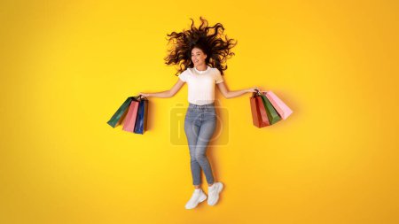 Foto de Feliz Shopaholic. Mujer joven sosteniendo coloridas bolsas de compras sonriendo a la cámara acostada en el fondo amarillo del estudio, vista superior. Comprador alegre Anuncio Gran oferta de ventas. Panorama - Imagen libre de derechos