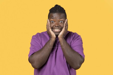 Téléchargez les photos : Omg, Wow Offre. Homme afro-américain émotionnel en T-shirt violet touchant la tête, regardant avec étonnement et choc à la caméra posant sur fond jaune, Studio Shot. Nouvelles étonnantes - en image libre de droit