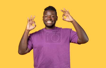 Foto de Retrato de un joven negro positivo mostrando un gesto aceptable en la cámara de pie sobre el fondo amarillo del estudio, señalando un signo de aprobación con ambas manos. Todo está bien Concepto - Imagen libre de derechos