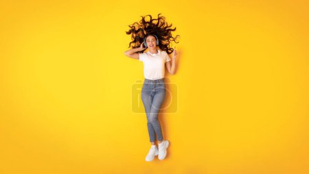 Photo for Digital Musical Fun. Excited Young Lady Listens To Music With Wireless Headphones, Points Finger Up And Enjoys Favorite Song Lying On Yellow Studio Background, Above View, Panorama - Royalty Free Image