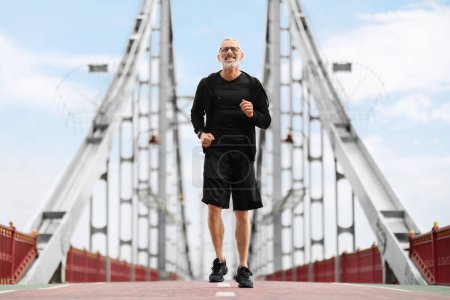 Téléchargez les photos : Grand-père homme senior courir en marchant pont, profiter de l'entraînement en plein air. Joyeux sportif motivé âgé jogging dans la rue par une journée chaude ensoleillée. Mode de vie sain pour les aînés concept - en image libre de droit