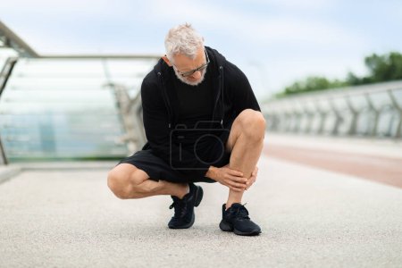 Téléchargez les photos : Homme âgé portant des vêtements de sport noirs jogging extérieur par pont, accroupi et toucher sa jambe, sportif à la retraite souffrant d'attelle du tibia, a été blessé pendant l'entraînement, copier l'espace - en image libre de droit