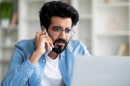 Photo for Confused Indian Man Looking At Laptop Screen And Talking On Cellphone, Young Eastern Male Suffering Problems With Computer, Reading Unpreasant Email, Having Mobile Phone Conversation, Closeup Shot - Royalty Free Image