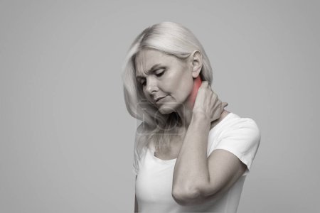 Photo for Unhappy mature woman feeling exhausted and suffering from neck pain, touching inflamed red zone, beautiful senior female having neckache, standing isolated on studio background, black and white photo - Royalty Free Image