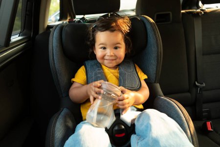 Photo for Baby Car Adventures. Smiling Asian Child Girl Posing Seated in Safe Toddler Vehicle Seat with Fastened Safety Straps, Relaxing and Enjoying Comfortable Auto Ride, Holding Water Bottle - Royalty Free Image
