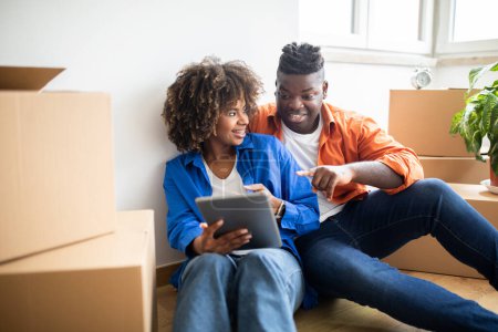 Foto de Cónyuges negros sonrientes que usan la tableta digital mientras que reubican a nuevo apartamento, pareja afroamericana feliz que se sienta en la habitación entre las cajas de cartón después de moverse, eligiendo muebles en línea - Imagen libre de derechos