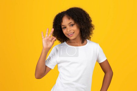 Téléchargez les photos : Joyeux adolescent élève noire en t-shirt blanc montre ok signe de la main, isolé sur fond jaune, studio. Étude Appruv, bonnes nouvelles, vente et retour à l'école, annonce et offre, éducation - en image libre de droit