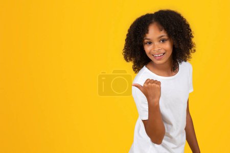 Téléchargez les photos : Adolescente élève noire positive en t-shirt blanc pointe du doigt sur l'espace vide, recommande étude, isolé sur fond jaune, studio. Vente approuve, école, annonce et offre, conseils éducatifs - en image libre de droit