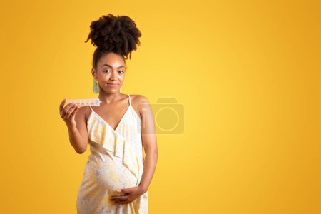 Téléchargez les photos : Joyeux millénaire noir dame enceinte toucher gros ventre, profiter de la grossesse, montrer des pilules de vitamines, isolé sur fond orange, studio. Publicité et offre, attente de l'enfant, maternité et soins de santé - en image libre de droit