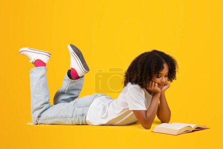 Photo for Smiling teenage black pupil girl in white t-shirt reads book, lies on floor, isolated on yellow background, studio. Study and back to school, ad and offer, hobby at free time, education, lifestyle - Royalty Free Image