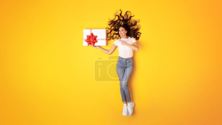 Téléchargez les photos : Surprise des fêtes. Happy Lady pointant vers la boîte de cadeau enveloppée sur la main, montrant son cadeau d'anniversaire et souriant à la caméra posant sur fond jaune dans Studio, Panorama avec espace libre, Vue du dessus - en image libre de droit