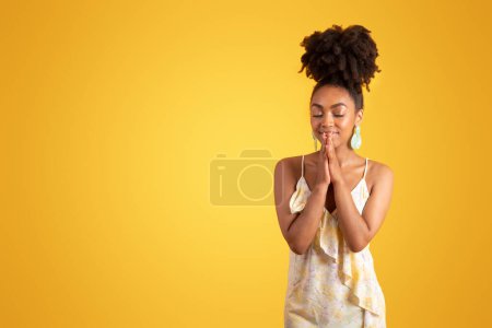 Foto de Señora negra milenaria positiva en vestido con los ojos cerrados rezando con las manos aisladas sobre fondo naranja, estudio. Gracias signo, petición, oración pedir deseo, anuncio y oferta, fe, esperanza emociones humanas - Imagen libre de derechos