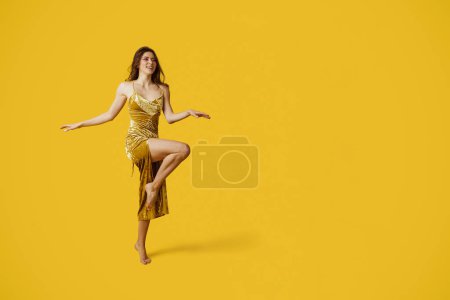 Foto de Tiro de longitud completa de la señora de moda en vestido elegante de oro posando sobre fondo de estudio amarillo, espacio libre. Emocionado mujer joven coqueta en traje de fiesta de moda - Imagen libre de derechos