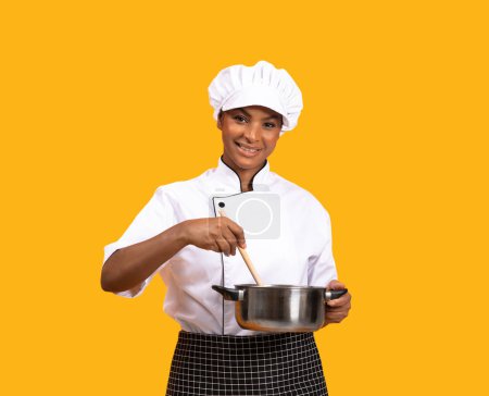 Photo for Beautiful Black Chef Woman With Spatula Stirring Food In Saucepan And Smiling At Camera, Happy Young African American Cook Lady Standing Isolated Over Yellow Studio Background, Copy Space - Royalty Free Image