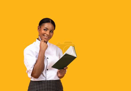 Photo for Black happy chef woman wearing uniform holding notepad and pen, smiling african american cook female noting recipes, writing cookbook while standing isolated on yellow studio background, copy space - Royalty Free Image