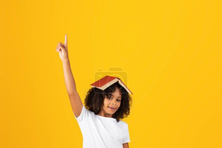 Foto de Muchacha adolescente adolescente adolescente en camiseta blanca con libro en la cabeza, levanta la mano, se divierte, aislado en el fondo amarillo, estudio. Hobby, estudiar y volver a la escuela, anuncio y oferta, educación, ganar - Imagen libre de derechos