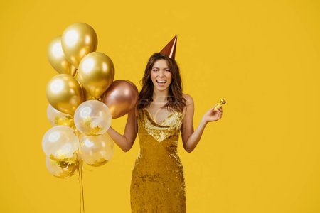 Téléchargez les photos : Dame satisfaite en robe avec des ballons festifs, casquette d'anniversaire et ventilateur de fête célébrant occasion spéciale, posant sur fond de studio jaune. Femme heureuse s'amuser en vacances - en image libre de droit