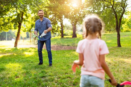 Téléchargez les photos : Vue arrière d'un homme d'âge moyen tenant une raquette de badminton et une grosse navette tout en jouant avec sa fille à l'extérieur dans le parc le jour de l'été. passe-temps et valeurs familiales - en image libre de droit