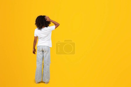 Foto de Chica negra adolescente pensativa en camiseta blanca se rasca la cabeza con la mano, aislado sobre fondo amarillo, estudio, longitud completa, espalda. Pregunta en el estudio, elegir y volver a la escuela, anuncio y oferta, educación - Imagen libre de derechos