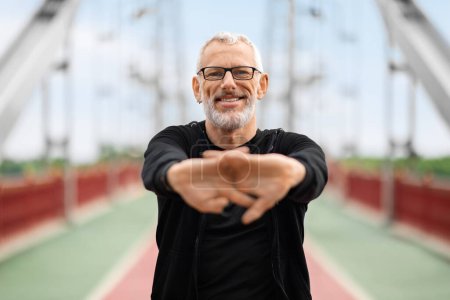 Photo for Happy motivated well-fit senior grey-haired sportsman wearing black sportswear exercising stretching hands before jogging on city walking bridge. Retired man have outdoor workout, copy space - Royalty Free Image