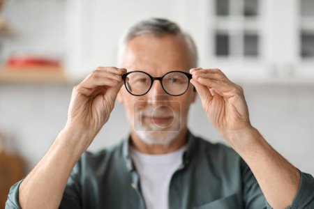Photo for Eye Vision Concept. Smiling Senior Man Looking At Camera Through Eyeglasses In His Hands, Handsome Elderly Gentleman Trying New Glasses While Standing At Home Interior, Selective Focus - Royalty Free Image