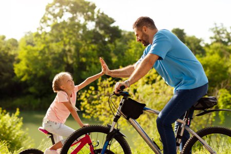 Téléchargez les photos : Père et sa fille donnant cinq tout en faisant du vélo dans un parc ensoleillé, profiter de passer du temps libre ensemble le week-end, vue latérale. Concept des valeurs familiales - en image libre de droit