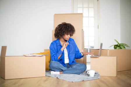 Foto de Mujer joven negra pensativa sentada entre cajas de cartón y usando el ordenador portátil, mujer afroamericana milenaria que ordena los muebles para el nuevo apartamento y hace la lista de comprobación de necesidades, espacio de copia - Imagen libre de derechos