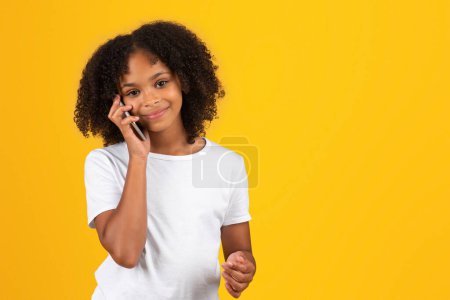 Photo for Happy teenage black pupil girl in white t-shirt calling by phone, isolated on yellow background, studio. Communication, study, good news and back to school, ad and offer, lifestyle education - Royalty Free Image