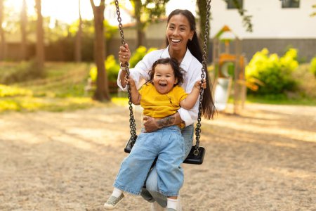 Foto de Playground Joy. Asiática madre y bebé niña balanceándose en los columpios, lleno de alegría mientras se divierten en el parque de atracciones al aire libre. Momentos reconfortantes de vínculo entre madre e hija - Imagen libre de derechos