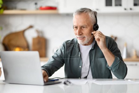 Photo for Smiling senior man sitting in kitchen, using modern laptop and headset, handsome elderly gentleman having video call with tutor, attending video conference or webinar, study or working online - Royalty Free Image