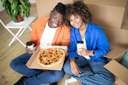 Photo for Cheerful black spouses eating pizza and drinking takeaway cofee in new home, happy african american couple having lunch break while moving apartment, relaxing on floor among cardboard boxes, closeup - Royalty Free Image