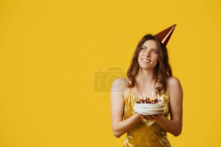 Téléchargez les photos : Femme rêveuse satisfaite dans le chapeau faire vœu et tenir gâteau avec des bougies, en regardant l'espace libre isolé sur fond jaune. Rêve, annonce et offre, fête d'anniversaire - en image libre de droit
