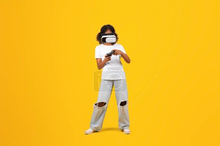 Téléchargez les photos : Joyeux adolescent fille noire gamer en t-shirt blanc, lunettes VR avec joystick profiter du temps libre, jouer à un jeu en ligne, isolé sur fond jaune, studio, pleine longueur. Fun, divertissement - en image libre de droit