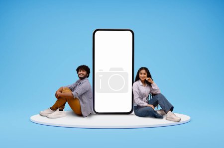 Photo for Cute cheerful smiling young eastern couple man and woman sitting on round platform by big phone with white blank screen, blue studio background, mockup blank space for mobile app, collage - Royalty Free Image