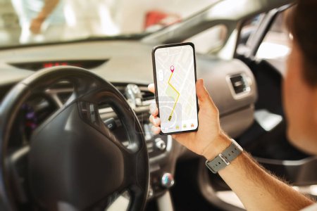 Téléchargez les photos : Homme conducteur assis à l'avant assis dans la voiture, tenant le téléphone mobile avec carte GPS navigation sur l'écran du gadget, gars méconnaissable vérifier la destination de l'emplacement, voyageant seul en auto - en image libre de droit
