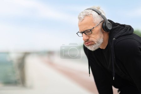 Photo for Closeup of tired exhausted senior man take break, catch heavy breath, elderly sportsman training jogging outdoors, using wireless headphones, listening to music, copy space - Royalty Free Image