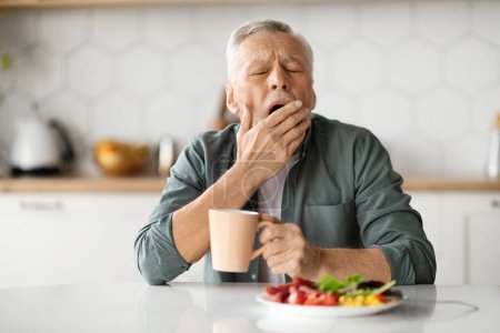 Übermäßige Tagschläfrigkeit. Müder Senior gähnt am Tisch in der Küche, älterer Herr schläfrig beim Mittagessen zu Hause, Mund mit der Hand bedeckt, Hypersomnie-Störung