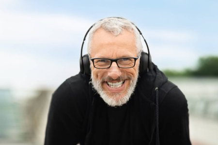 Photo for Closeup of happy grey-haired handsome senior sportsman wearing black sportswear and wireless headphones have morning workout routine outdoor, retired man rest after jogging, smiling at camera - Royalty Free Image