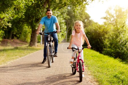 Photo for Family pastime concept. Happy middle aged father and his pretty daughter riding bicycles together, walking and spending time in the park on a sunny day - Royalty Free Image
