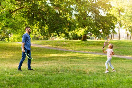 Téléchargez les photos : Un passe-temps familial. Vue latérale de papa jouant au badminton avec sa fille, tenant des raquettes et utilisant une grande navette, passant du temps à l'extérieur dans le parc le jour de l'été - en image libre de droit